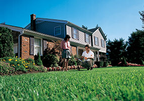 A lawn tech employee crouching over grass in front of a home speaking with a woman.