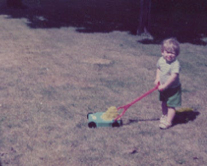 Rick Longnecker as a young child pushing a toy lawnmower on grass